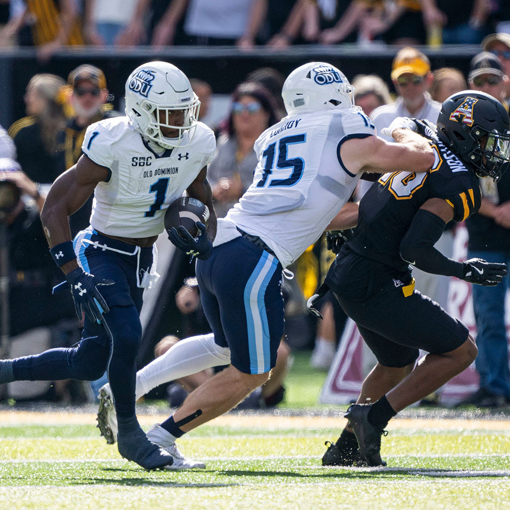 Football game action with a player in a white Old Dominion uniform holding the ball, while another in black attempts a tackle. Tension and focus are evident.